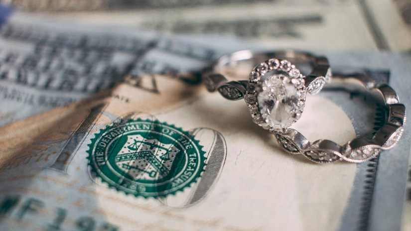 In a macro photo, two rings rest against a dollar bill, representing savings for an engagement ring budget.